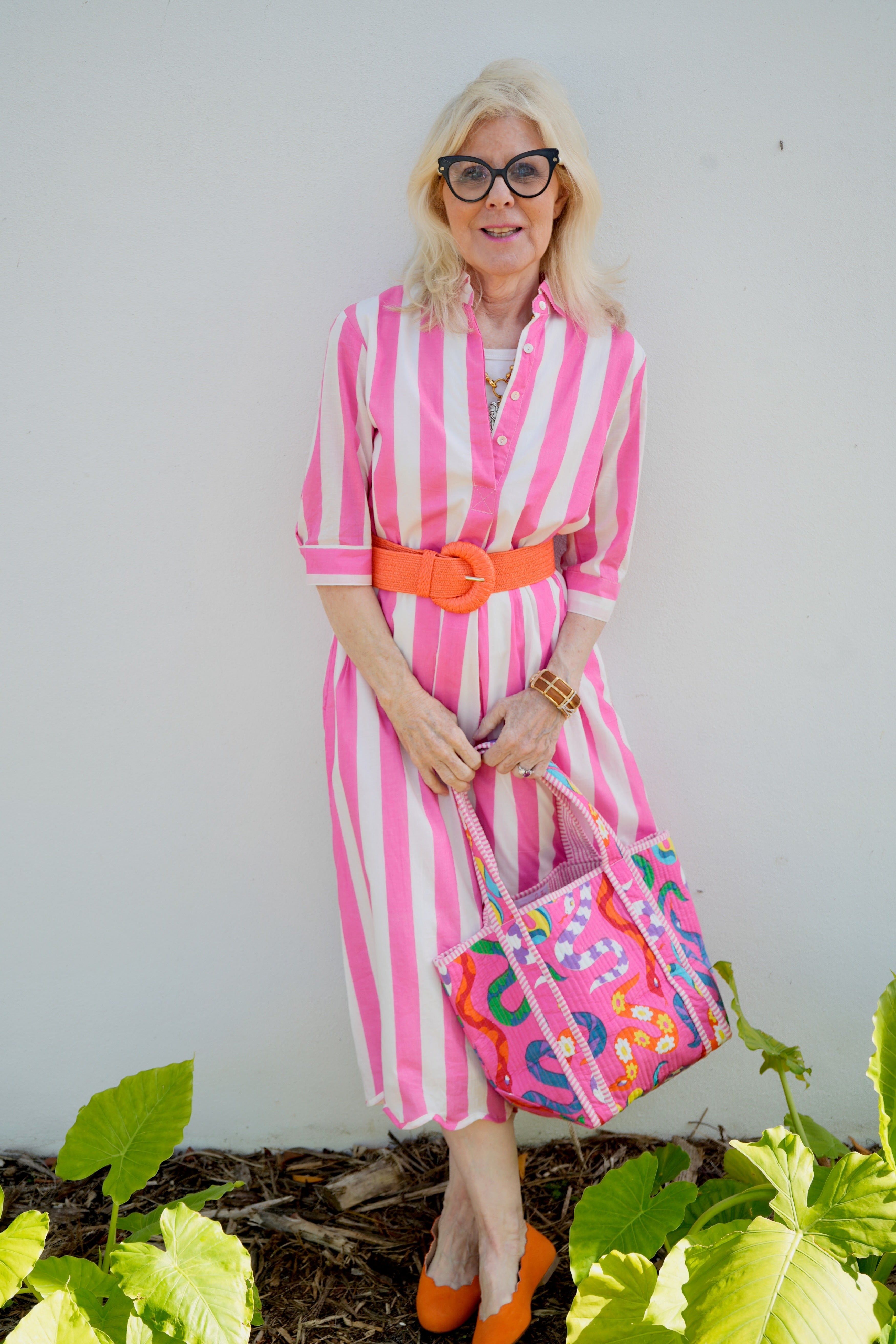 Woman in a pink and white striped dress with an orange belt, holding a colorful handbag, standing against a white wall with green plants.