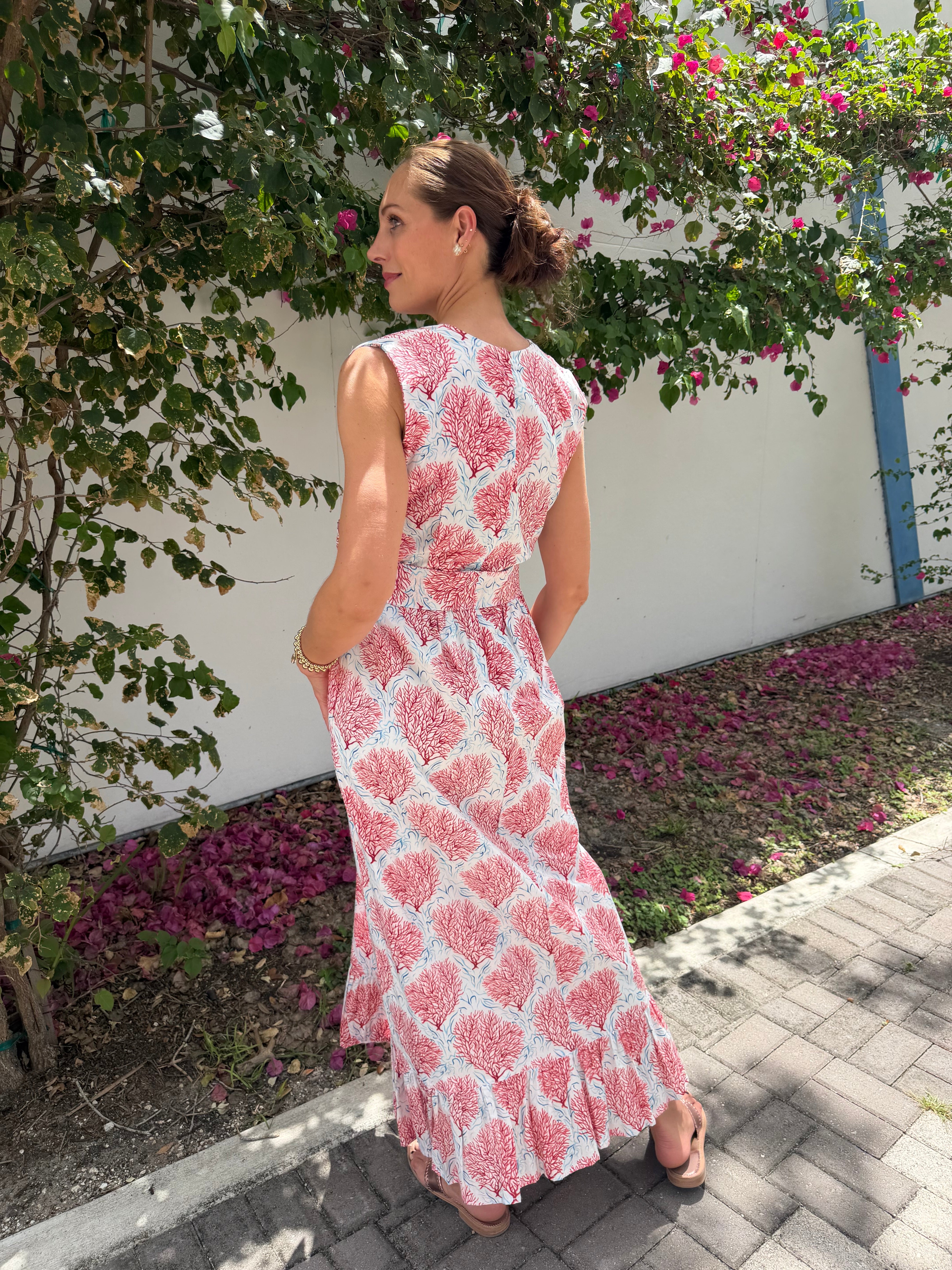 Woman in a pink floral dress standing in a garden with flowers and a white wall.