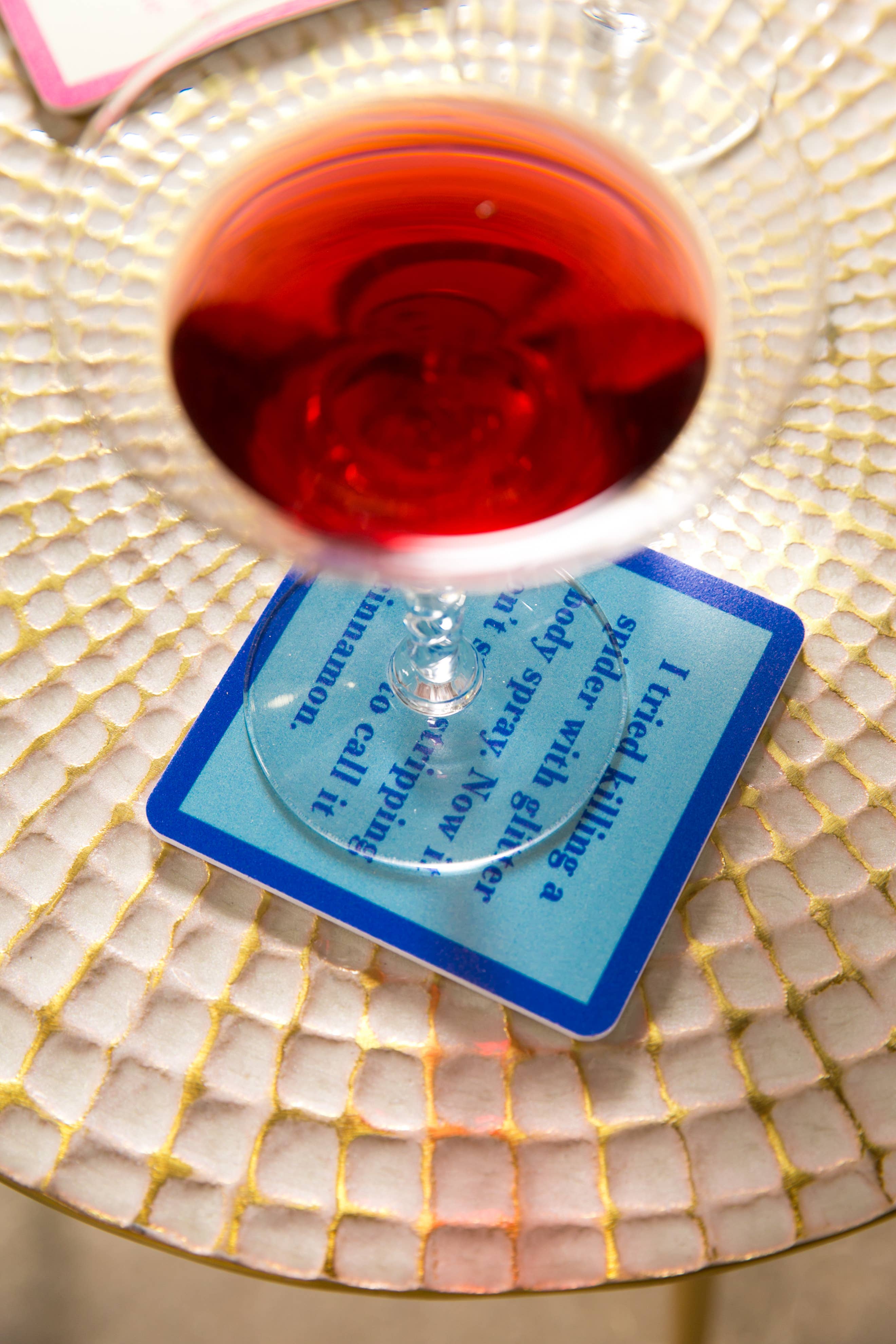 Cocktail glass with red liquid on a decorative coaster with text, placed on a mosaic-patterned surface.