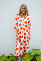 Woman wearing a dress with orange fruit pattern standing against a white wall with green plants.