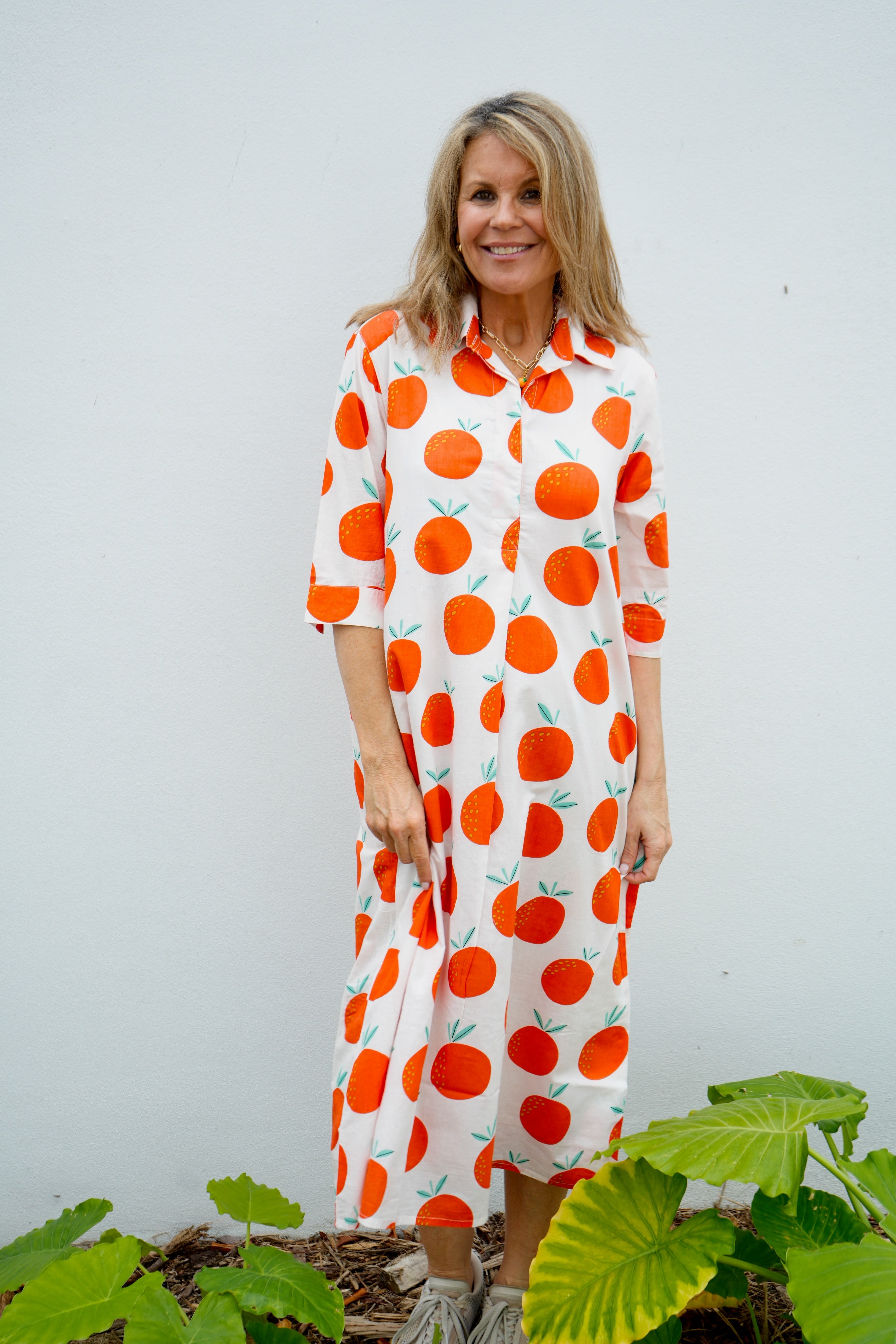 Woman wearing a dress with orange fruit pattern standing against a white wall with green plants.