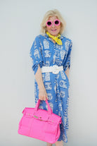Woman in a blue dress with white patterns, holding a pink bag, wearing pink sunglasses, and a yellow scarf against a plain background.