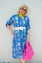 Person wearing a blue patterned dress with a white belt, holding a pink handbag against a plain background.