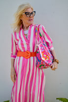 Woman in a pink and white striped dress with an orange belt, holding a colorful bag against a plain background.