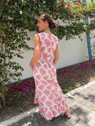 Woman in a pink floral dress standing in a garden with flowers and a white wall.